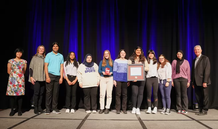 The 2023 Peace Medal Youth Award winners North Park Collegiate’s Student Anti-Racism Coalition (SARC) pose with their award and the presenters in front of a blue curtain backdrop on stage.