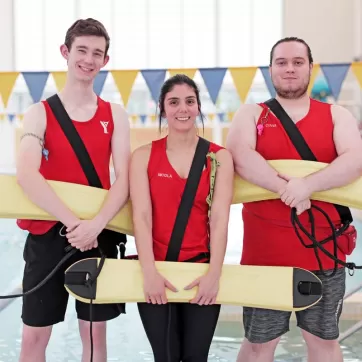 An image of two males and one female YMCA lifeguards smiling as they hold on to their safety equipment in front of a pool.