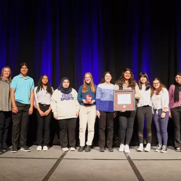 The 2023 Peace Medal Youth Award winners North Park Collegiate’s Student Anti-Racism Coalition (SARC) pose with their award and the presenters in front of a blue curtain backdrop on stage.
